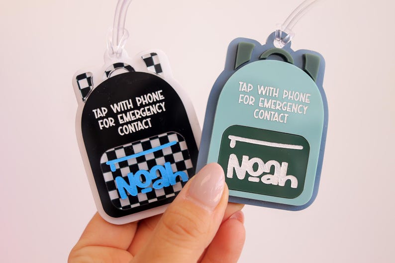 May include: Two backpack-shaped luggage tags, one black with a checkered pattern and one teal. Both tags have the text "TAP WITH PHONE FOR EMERGENCY CONTACT" and the name "Noah".