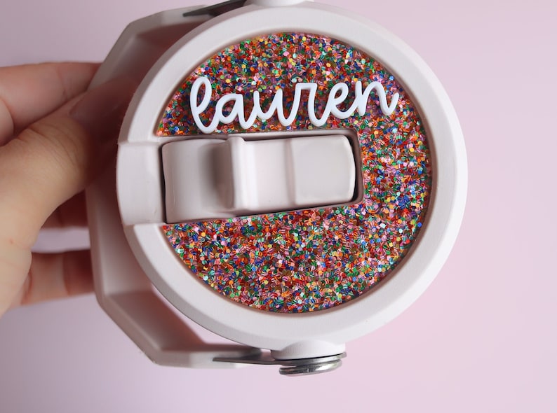 May include: A light pink water bottle with a white lid and a circular design filled with colorful glitter. The name "lauren" is written in white cursive across the glitter. The bottle has a white handle and a silver base.