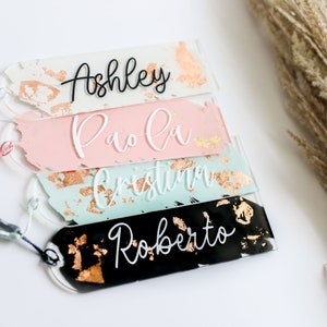 May include: Four acrylic bookmarks with gold foil accents and white lettering. The names Ashley, Paola, Christina, and Roberto are written on the bookmarks.