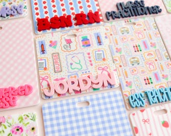May include: A collection of rectangular badge reels featuring various patterns including gingham, floral, and medical icons. Some reels display raised 3D lettering with names or titles like JORDYN, BSN RN, NURSE PRACTITIONER, REGISTERED NURSE, and ESS TEACHER.
