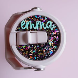May include: White water bottle lid with a flip-top closure and a handle. The lid features a circular design filled with iridescent glitter and the name "emma" in teal script. The background is a soft pink.