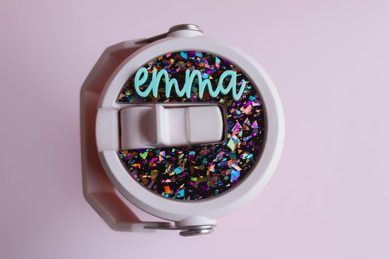 May include: White water bottle lid with a flip-top closure and a handle. The lid features a circular design filled with iridescent glitter and the name "emma" in teal script. The background is a soft pink.