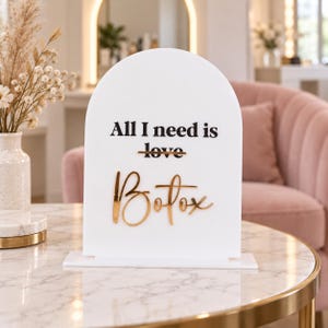 May include: A white arched acrylic sign featuring the black text All I need is love with the word love crossed out. Below this, the word Botox is written in a reflective gold script font. The sign stands on a rectangular white base.
