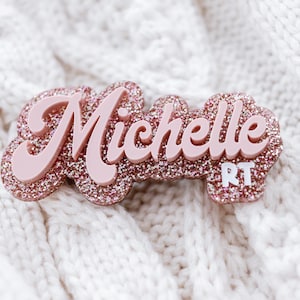 May include: A pink glitter name tag with the name "Michelle" in a cursive font and the letters "RT" in white.