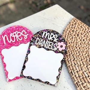 May include: Two decorative acrylic note boards. One is pink with the word "notes" in white script. The other is black with the words "mrs DANIELS" and a pink flower. Both have a white space for writing.