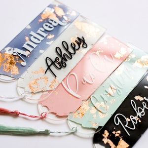 May include: Five personalized acrylic bookmarks with gold leaf accents and tassels. The bookmarks are in various colors, including blue, pink, green, and black. The names "Andrea", "Ashley", "Paul", "Olivia", and "Roberto" are written on the bookmarks.