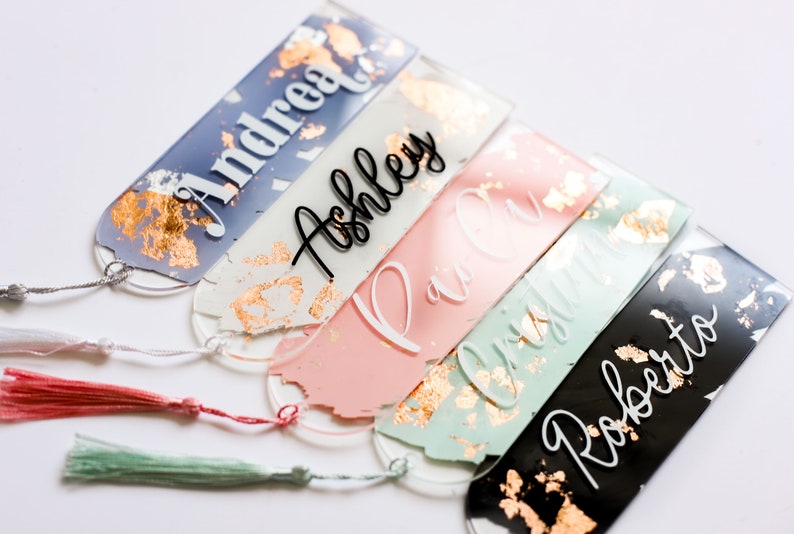 May include: Five personalized acrylic bookmarks with gold leaf accents and tassels. The bookmarks are in various colors, including blue, pink, green, and black. The names "Andrea", "Ashley", "Paul", "Olivia", and "Roberto" are written on the bookmarks.