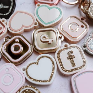May include: Assortment of colorful, glittery, and metallic charms featuring social media icons like Instagram, Facebook, and a heart. The charms are in various shapes, including squares, hearts, and circles.