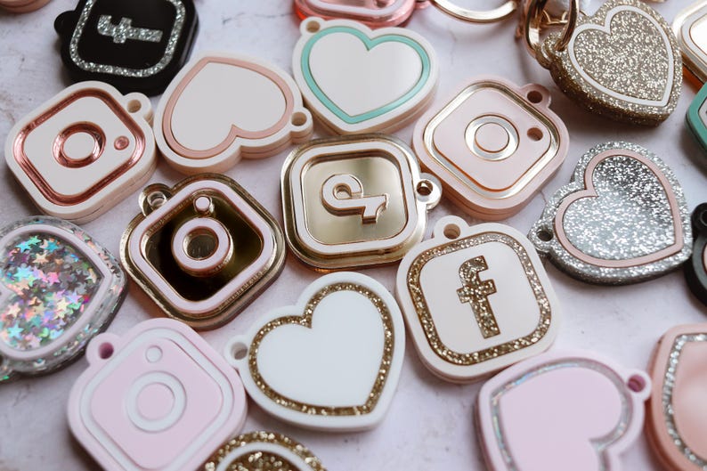 May include: Assortment of colorful, glittery, and metallic charms featuring social media icons like Instagram, Facebook, and a heart. The charms are in various shapes, including squares, hearts, and circles.