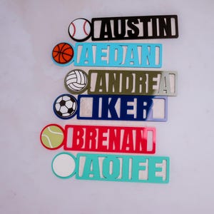 May include: A set of seven personalized name tags with different sports ball icons. The names are Austin,  Jedan, Andrea, Iker, Brenan, and Aoife. The tags are made of acrylic and come in a variety of colors.
