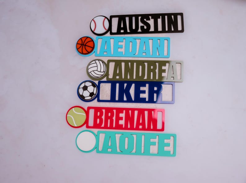 May include: A set of seven personalized name tags with different sports ball icons. The names are Austin,  Jedan, Andrea, Iker, Brenan, and Aoife. The tags are made of acrylic and come in a variety of colors.