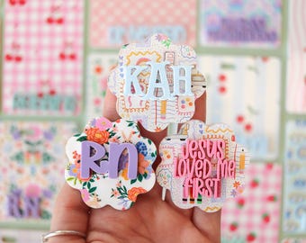 May include: Three scalloped-edge badge reels featuring decorative patterns. The top one displays light blue letters KAH. The bottom left features a floral print with purple letters RN. The bottom right has a doodle pattern with pink text Jesus loved me FIRST.