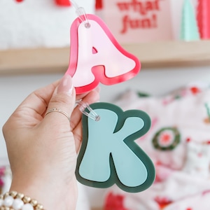 May include: A pink letter A and a teal letter K, both with a glossy finish, are strung together on a clear string. The letters are part of a banner and are being held by a hand.