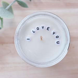 May include: A white candle in a clear glass jar, with the words "I LOVE YOU" spelled out in small, black letter beads. The candle sits on a light-colored wooden surface, with a sprig of greenery in the background.