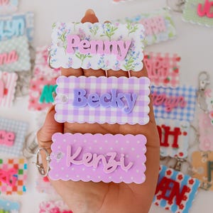 Op de afbeelding: Drie rechthoekige sleutelhangers met geschulpte randen en verhoogde 3D-namen. De bovenste sleutelhanger heeft een bloemenpatroon met de naam Penny, de middelste een paars ruitpatroon met de naam Becky en de onderste paarse stippen met de naam Kenzi.