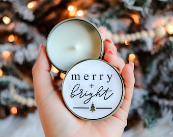 Puede incluir: Una pequeña vela redonda en una lata plateada, con un interior de cera blanca y una mecha. La tapa presenta las palabras "merry + bright" en escritura negra, con un pequeño gráfico de árbol de Navidad. La vela se sostiene en una mano.