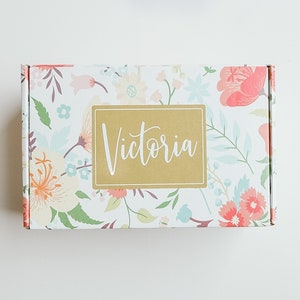 Puede incluir: Una caja blanca con un patrón floral y una etiqueta dorada que dice "Victoria".