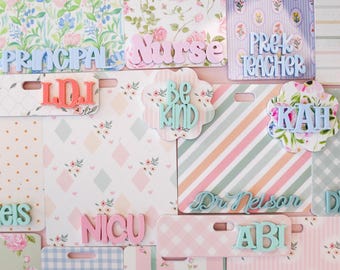 May include: A collection of rectangular and scalloped name badges featuring various floral, striped, and checkered patterns. The badges display raised 3D lettering in pastel colours with text including PRINCIPAL, Nurse, PREK TEACHER, LDI, be kind, KAH, NICU, Dr Nelson, ABI, and DRE.