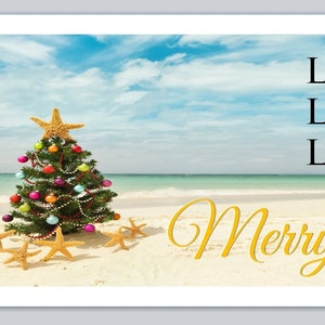 May include: A Christmas tree decorated with colorful ornaments and lights on a white sandy beach with blue sky and ocean in the background. The text "Merry Christmas" is written in gold script on the sand.