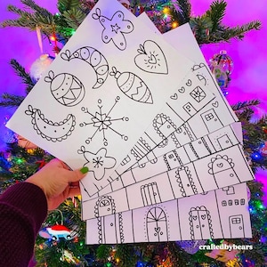 May include: A set of five printable Christmas colouring pages featuring simple black line drawings of ornaments, snowflakes, stars, hearts, and houses. The pages are stacked on top of each other and held in a hand against a background of a decorated Christmas tree.