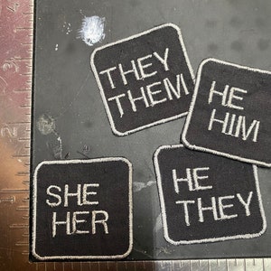 Può includere: Quattro toppe di tessuto nero con testo bianco. Le toppe recitano "They Them", "He Him", "She Her" e "He They".