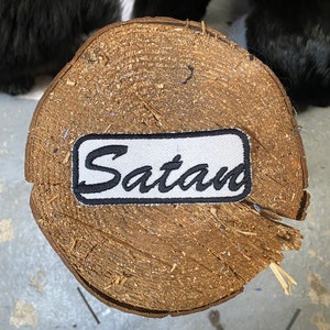 Può includere: Toppa rettangolare bianca con testo ricamato nero che recita "Satan" su una superficie di legno.
