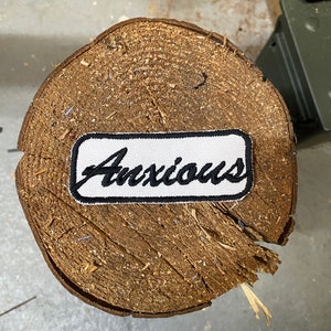 May include: White embroidered patch with black stitching and the word "Anxious" in a cursive font.