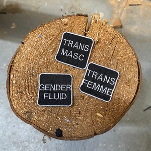 Op de afbeelding: Drie zwarte stoffen patches met witte tekst. De patches lezen "Trans Masc", "Trans Femme" en "Gender Fluid".