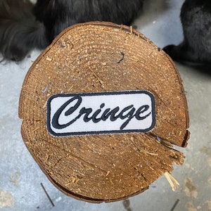 May include: White patch with black embroidered text that reads "Cringe" on a wooden surface.