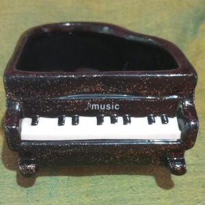 May include: A small, decorative ceramic planter shaped like a grand piano. The planter is dark brown with a white keyboard and the word "music" printed on the front. The interior is black.