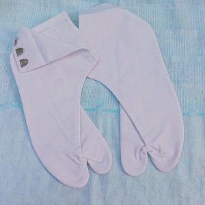 Può includere: Un paio di calzini tabi bianchi, tradizionali calzini giapponesi a punta divisa, sono esposti su una superficie strutturata azzurra. Un calzino ha un risvolto piegato fissato con due piccoli bottoni color argento.