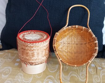 May include: Two woven bamboo baskets. One is a round, lidded container with a red trim and a red string handle. The other is a round basket with a handle. Both are made of light brown woven bamboo.