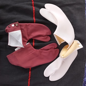 Può includere: Due paia di calze tabi, un paio è bianco e l'altro è bordeaux. Le calze sono su una superficie di tessuto nero.