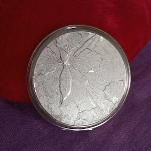 May include: A silver compact mirror with a floral design. The mirror has a raised, textured design of a flower with leaves.