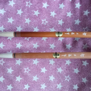 Könnte beinhalten: Zwei Kalligraphiepinsel mit hellbraunen Holzgriffen und weißen Borsten werden auf einer rosa Oberfläche mit weißen Sternmustern gezeigt. Die Pinsel haben goldene Etiketten und asiatische Schriftzeichen aufgedruckt. Ein Pinsel hat eine schwarze Schlaufe.