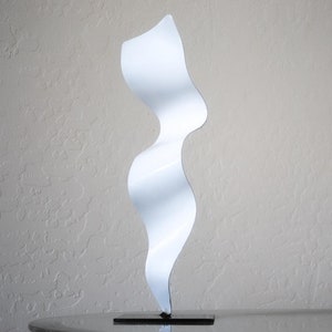 May include: White abstract metal sculpture with a wavy, organic shape. The sculpture is on a black base and is standing on a table.