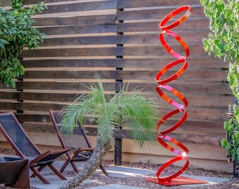 Op de afbeelding: Een rode metalen sculptuur in de vorm van een spiraal, die in een tuin staat. De sculptuur is gemaakt van meerdere overlappende cirkels, wat een dynamisch en modern ontwerp creëert. De sculptuur staat op een kleine, vierkante basis.