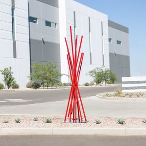 May include: A red metal sculpture resembling a tree with branches extending upwards. The sculpture is in front of a modern building with gray and white panels.
