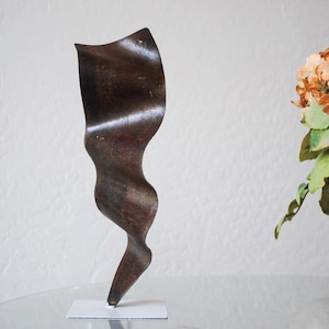 Op de afbeelding: Een donkerbruin metalen sculptuur met een golvend, abstract ontwerp. Het sculptuur staat op een witte basis.