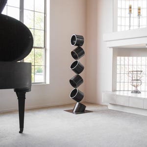 May include: A black metal sculpture of stacked cylinders, resembling a tower, stands on a white floor in a modern home. The sculpture is made of six cylinders, each slightly offset from the one below it, creating a dynamic and abstract form.
