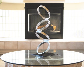 May include: A silver metal sculpture of four stacked rings, each ring slightly larger than the one above it, on a black base. The sculpture is on a glass-top table in front of a fireplace.