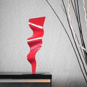 Op de afbeelding: Een rood metalen beeldhouwwerk met een golvend, abstract ontwerp. Het beeldhouwwerk staat op een zwart oppervlak.