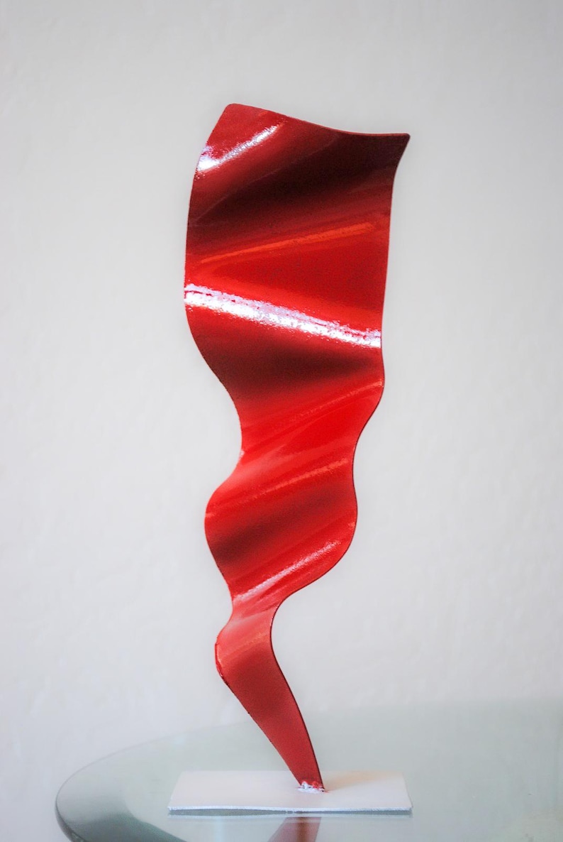 May include: A red abstract metal sculpture with a wavy, flowing design. The sculpture is standing on a white base.