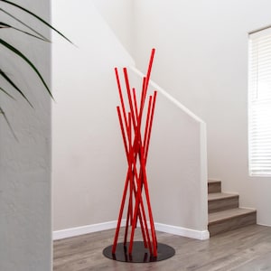 May include: A red metal coat rack with a circular base, standing in a white room with a wooden staircase in the background.