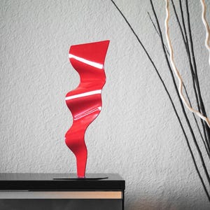 May include: A red metal sculpture with a wavy, abstract design. The sculpture is standing on a black surface.