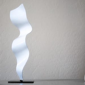 Op de afbeelding: Witte abstracte metalen sculptuur met een golvend, vloeiend ontwerp. Het beeld staat op een zwarte basis.