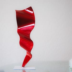 May include: A red metal sculpture with a wavy, abstract design. The sculpture is on a white base and is about 12 inches tall.