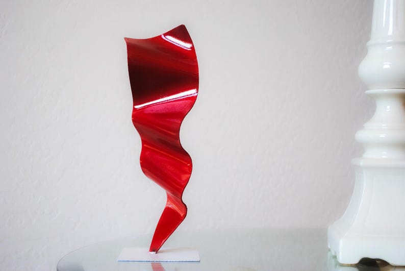 May include: A red metal sculpture with a wavy, abstract design. The sculpture is on a white base and is about 12 inches tall.