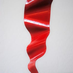 Op de afbeelding: Een abstract metalen beeldhouwwerk in rood met een golvend, vloeiend ontwerp. Het beeldhouwwerk staat op een witte basis.