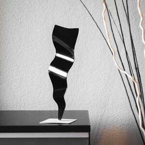 May include: A black metal sculpture with a wavy, abstract design. The sculpture is standing on a small, square base.